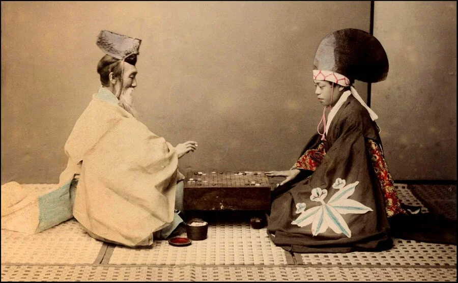 A primeira imagem mostra duas pessoas sentadas no chão, frente a frente, com um tabuleiro de Go entre elas. Uma pessoa está vestida com trajes tradicionais japoneses, com um robe branco e um chapéu alto e pontudo. A outra pessoa também está em vestimentas tradicionais, com um kimono preto adornado com um grande símbolo de flor branca e um grande laço na cabeça. Ambos estão concentrados no jogo, imersos na estratégia.