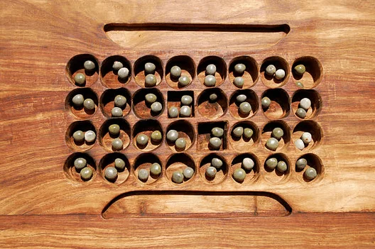 Um tabuleiro de Mancala feito de madeira polida, com duas fileiras de cavidades arredondadas onde as sementes ou pedras são colocadas e movimentadas durante o jogo. O tabuleiro tem uma coloração rica e quente e exibe uma forma retangular com uma depressão central que serve como depósito para as peças capturadas.