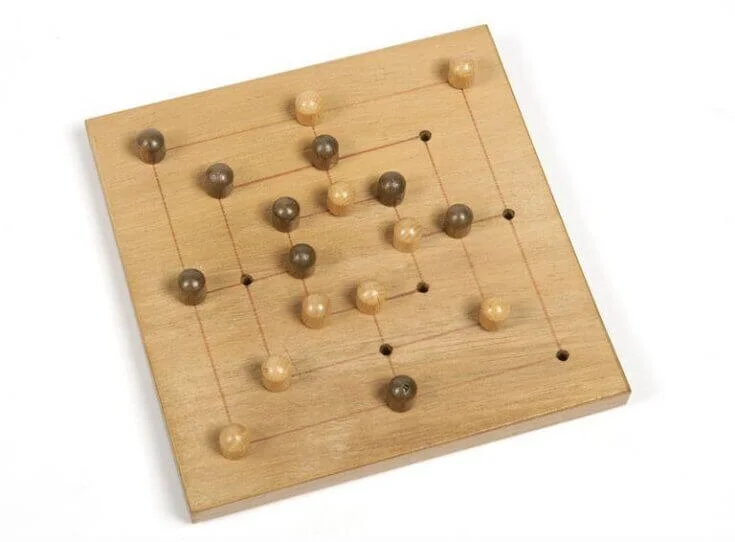 Um tabuleiro de Nine Men's Morris, feito de madeira clara com linhas gravadas conectando pontos onde as peças podem ser colocadas. O tabuleiro é quadrado, com um sistema de linhas que formam três quadrados concêntricos e linhas conectando os meios de cada lado. Peças esféricas em dois tons distintos, claras e escuras, estão organizadas em formação de jogo no tabuleiro.